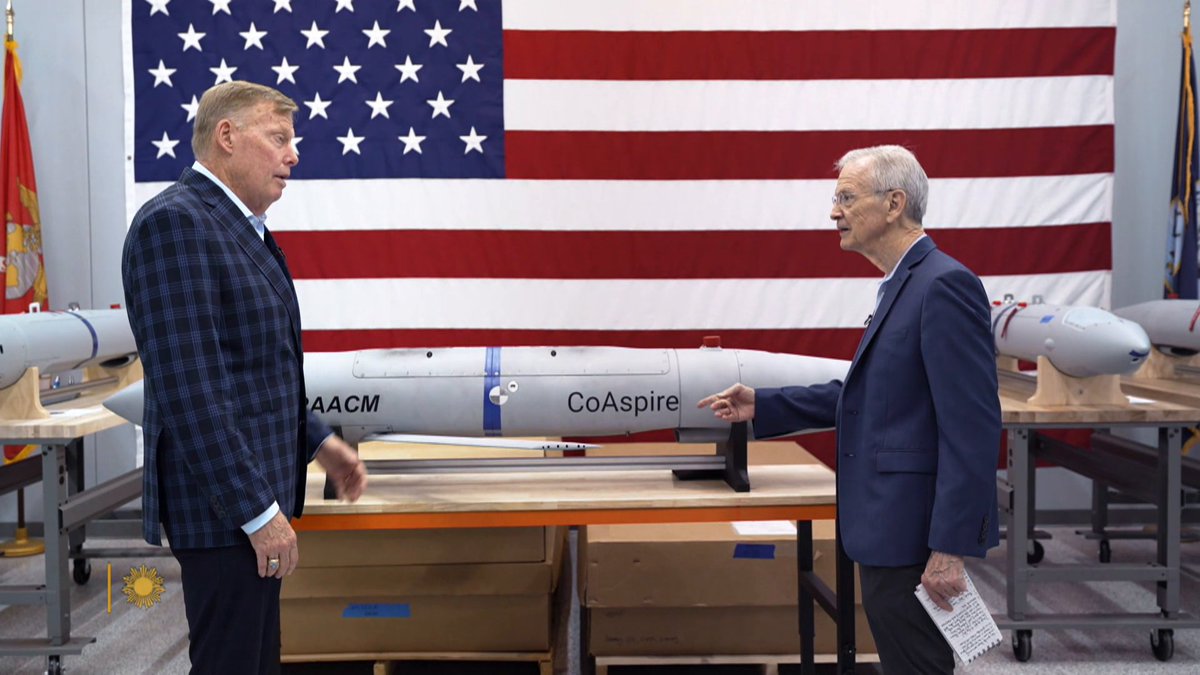 Two men in suits stand in front of a large American flag, discussing a missile labeled "AACM CoAspire" displayed on a table, highlighting Camden, Arkansas's role in producing critical defense munitions for American military forces.