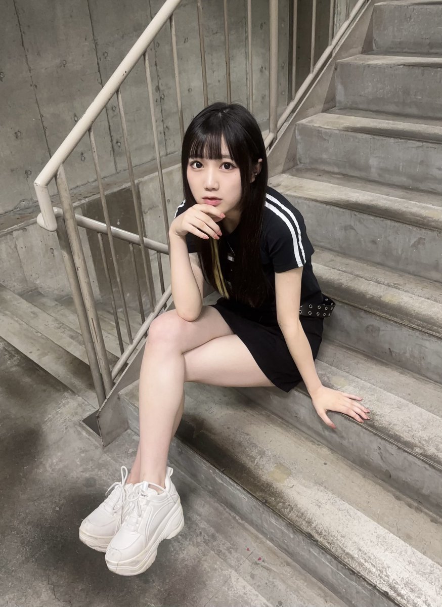 コンクリート打ちっぱなしの階段に座っている橘めい
を正面やや上から撮影した他撮り写真です。

黒い半袖トップスには肩から袖にかけて白いラインが2本入っており、黒いスカートを合わせています。
首元には鍵モチーフのペンダントをつけています。
腰にはシルバーの金具がついた黒いベルトを巻いています。
白い厚底タイプのスニーカーを履いています。

階段の途中に横向きに腰掛け、片方の脚を前方へ伸ばし、もう片方の脚を曲げています。片手は階段に置き、もう片方の手は顎の近くに添えています。

カメラを見つめており、お澄まし顔です。

背景はコンクリート壁と階段、金属製の手すりがあり、全体的にグレーを基調としたシンプルな空間です。