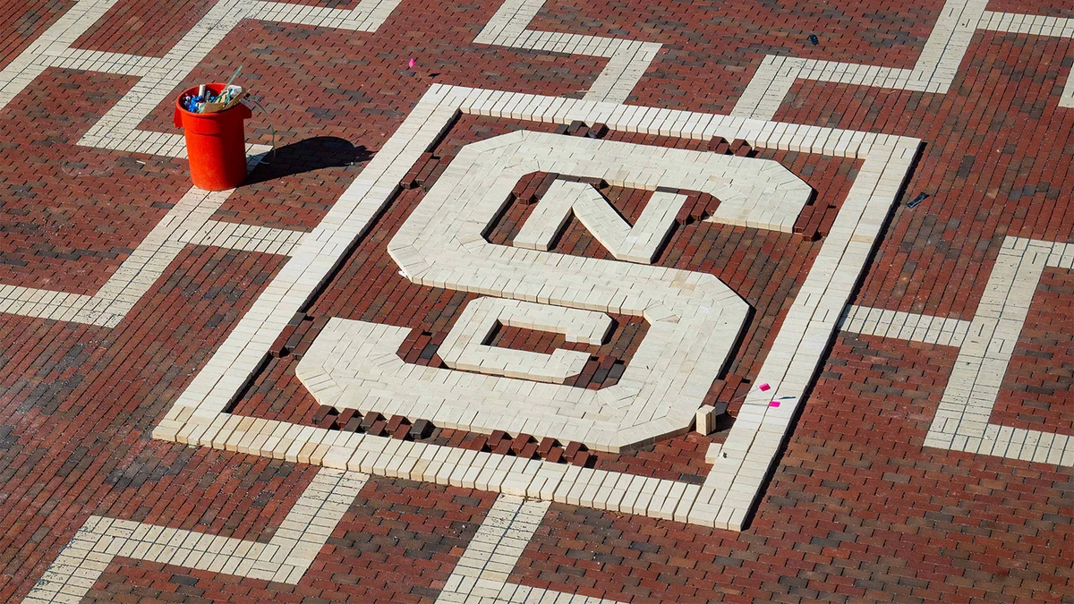 A large white brick design of the letters 'N' and 'C' intertwined on a red brick pavement with a red bucket of tools nearby.