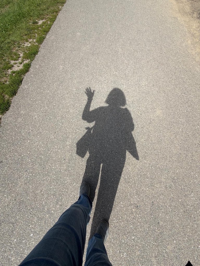 It’s my shadow on the ground. In the foreground are my actual jeans and trainers and then my shadow on the path. There’s grass off to the side. I have two bags. This often happens if I take a small handbag. I end up with a canvas bag for all of the rest of the stuff. My shadow hair is a bob and my shadow hand is waving.