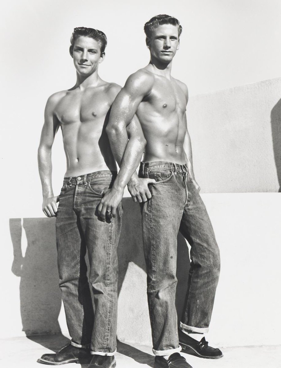 viaSargedna's tweet photo. This photograph has the unmistakable aesthetic of Bob Mizer and the Athletic Model Guild (AMG) in the 1950s: an idealised vision of youthful American masculinity in which physical fitness, informality, camaraderie and a certain carefully controlled sensuality are brought together.
The two models are posed outdoors against a stark, sunlit architectural setting. Their bare torsos and rolled-up denim jeans create a deliberately uncomplicated, working-class image of masculinity—more young men at leisure than classical studio models. Yet the pose is anything but accidental. Both figures stand upright and confidently, their bodies carefully presented to show the broad shoulders, narrow waists, developed chests and arms that were central to Mizer's physique aesthetic.

The composition is particularly effective because of the overlapping figures. The nearer model dominates the picture while his companion stands immediately beside him, producing a compact arrangement of two bodies rather than two isolated portraits. Their similar clothing, hairstyles and physiques reinforce the suggestion of youthful male companionship, while the slight differences in expression and stance prevent the image from becoming mechanically symmetrical.

The hard California sunlight is important aesthetically. It throws crisp shadows across the white wall and gives the bodies a sculptural quality, almost recalling the strong modelling of light found in classical sculpture. The models' smooth, oiled skin contrasts with the rough texture of the denim, creating one of Mizer's characteristic visual oppositions: the perfected male body against ordinary American clothing and surroundings.

There is also an intentional tension between the photograph's ostensibly wholesome subject and its sensual presentation of the male body. AMG photographs were frequently framed as studies of athleticism, health and physical culture, but Mizer understood very well how posture, proximity, exposed torsos and the emphasis on musculature could create an image with considerable homoerotic appeal. Here the aesthetic remains restrained: there is no overt sexual action, but the physical attractiveness of the two men is plainly the principal visual subject.

In that sense, the photograph presents a quintessential Mizer fantasy of the 1950s: the handsome, healthy, self-assured young American male—casual, athletic, sun-drenched and seemingly unconcerned with being looked at—while the photograph itself makes that body the object of admiration.