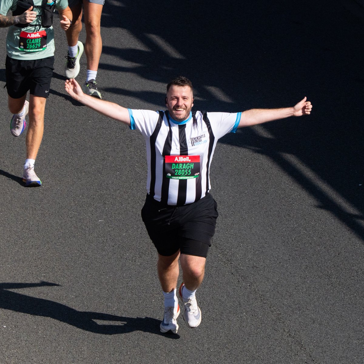 A person running in a striped sports jersey with the name "Darragh" and number "28055" visible, arms outstretched, on a sunny roadway. Other runners are partially visible in the background.