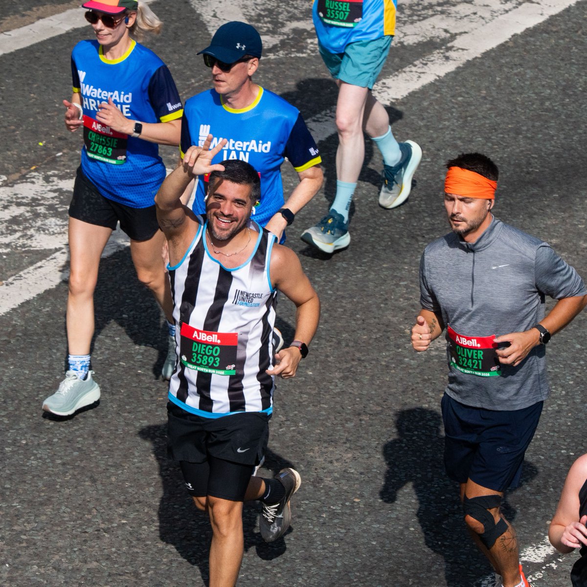 Group of runners in a marathon, with some wearing "WaterAid" shirts. A participant in a black and white striped shirt is smiling and waving at the camera. The race number and branding "ABell" are visible on event bibs.