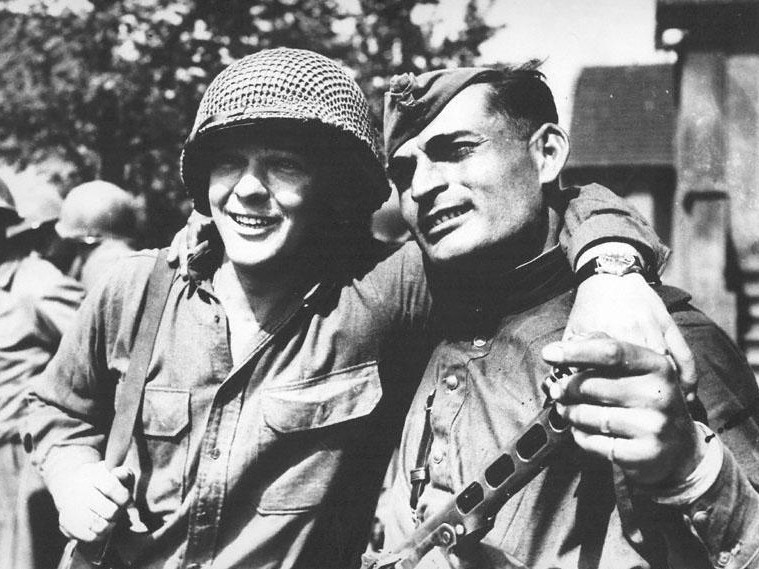 Two smiling soldiers from World War II, one wearing a net-covered helmet and the other a cap, stand closely together with arms around each other. The scene suggests camaraderie and relief. They are outdoors with trees and a building visible in the background.