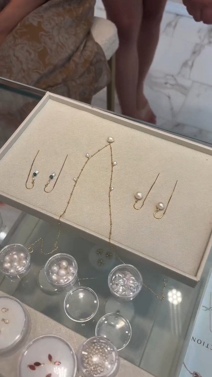 A jewelry display tray featuring pearl and gemstone earrings along with a gold chain necklace, with small containers of additional pearls visible on a glass surface.