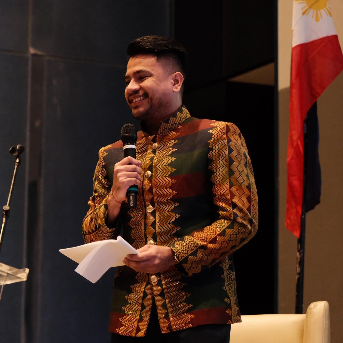 Mohammad speaks into a microphone while smiling and holding papers and wears a traditional formal jacket.
