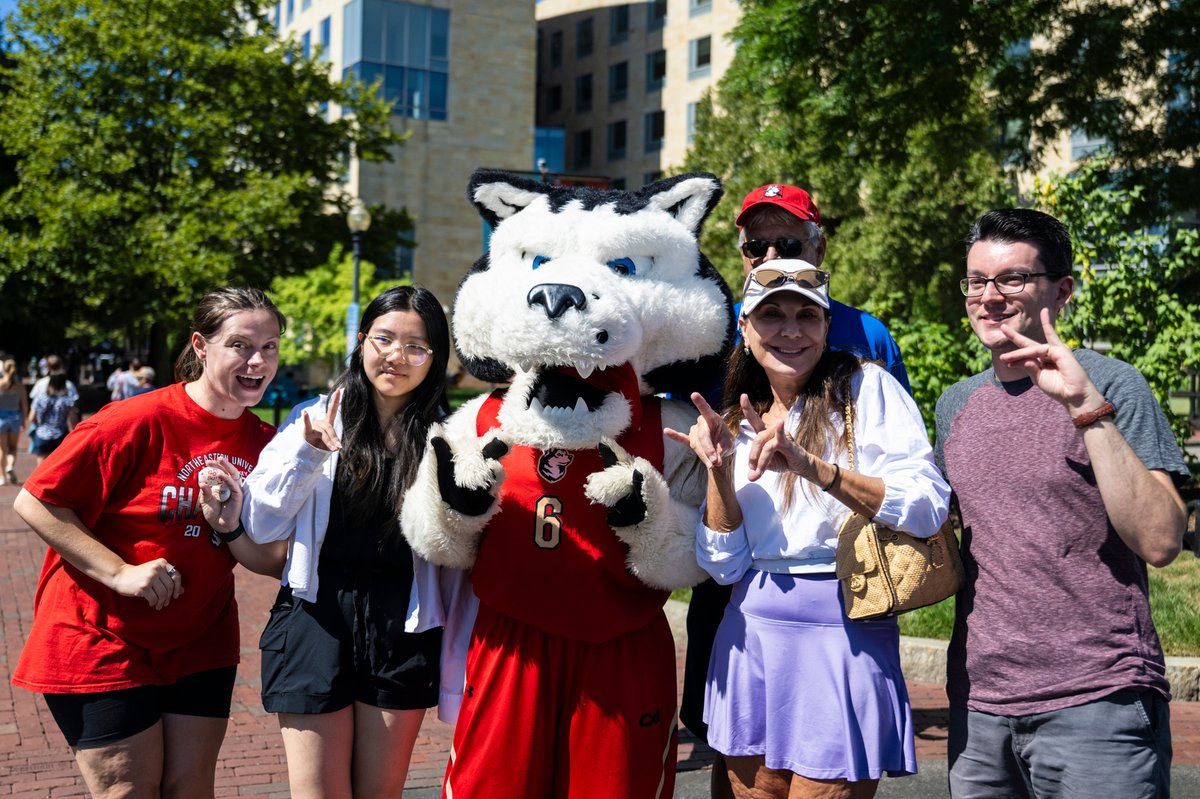 Five smiling people pose with a husky mascot in a red #6 jersey (CA visible). A woman in a red \"Northeastern Univ. Champ 20\" shirt, another in a white shirt, and a man in glasses make peace signs. Sunny outdoor setting with brick ground, trees & buildings.
