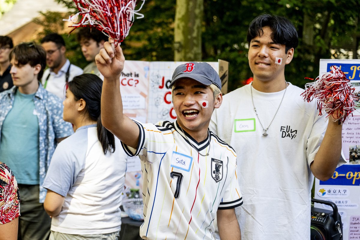 Two young men smile and cheer, holding red and white pom-poms. Both have Japanese flag stickers on their cheeks. Info boards visible in background.