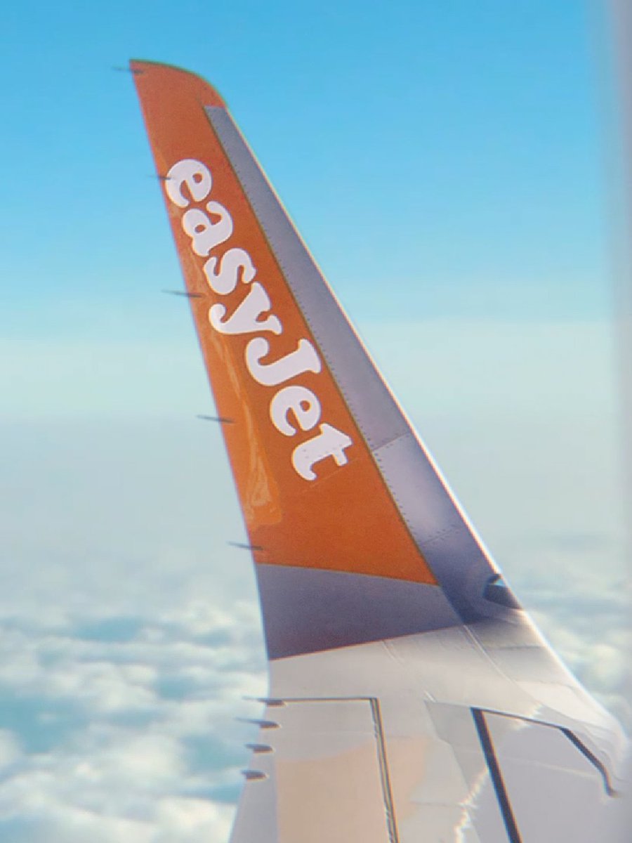The orange winglet of an easyJet plane with the brand name visible, seen from a window above a layer of white clouds.