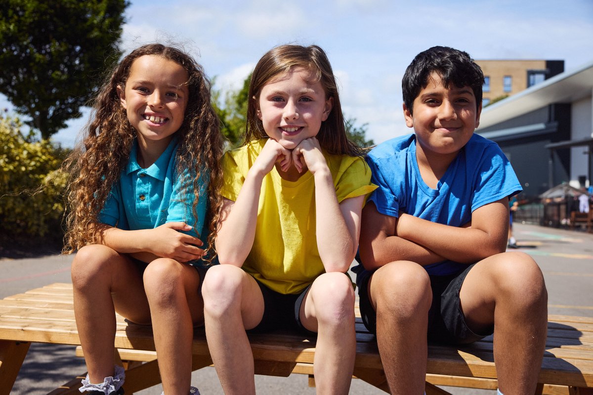 Three children sat on a wooden bench outdoors, smiling and enjoying a sunny day at school.