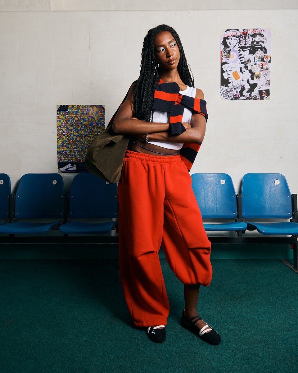 An image of a person wearing a pair of black and white PUMA Speedcat Ballet shoes with orange jogging bottoms and white top whilst standing in front of a white wall lined with blue chairs.