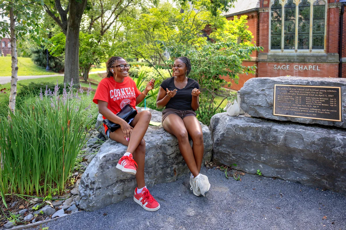Two people are sitting on a stone ledge near Cornell's Sage Chapel. One is wearing a red Cornell t-shirt. They are conversing and surrounded by greenery.