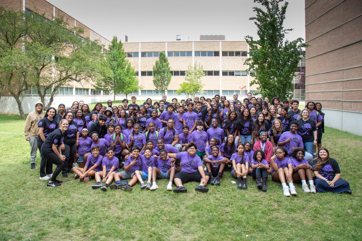 A large group Oakland University Math Corps attendees wearing matching purple shirts pose outdoors on a grassy area between buildings. The setting appears to be a campus or school environment, with trees and buildings in the background.