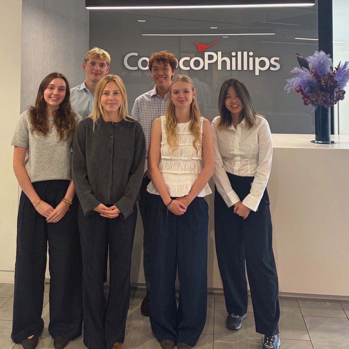 A group of six people standing in front of a reception desk with a ConocoPhillips sign visible on the wall. They are all dressed in smart-casual clothing, and there is a floral arrangement on the desk.