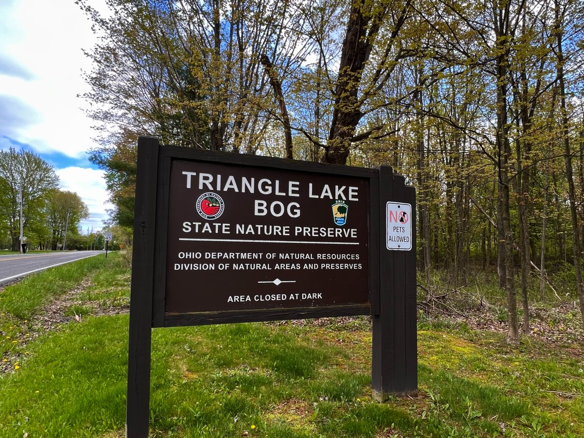 Sign for Triangle Lake Bog State Nature Preserve, operated by the Ohio Department of Natural Resources. It states the area is closed at dark. A "No Pets Allowed" sign is also visible. Surrounded by trees and a road on the left.