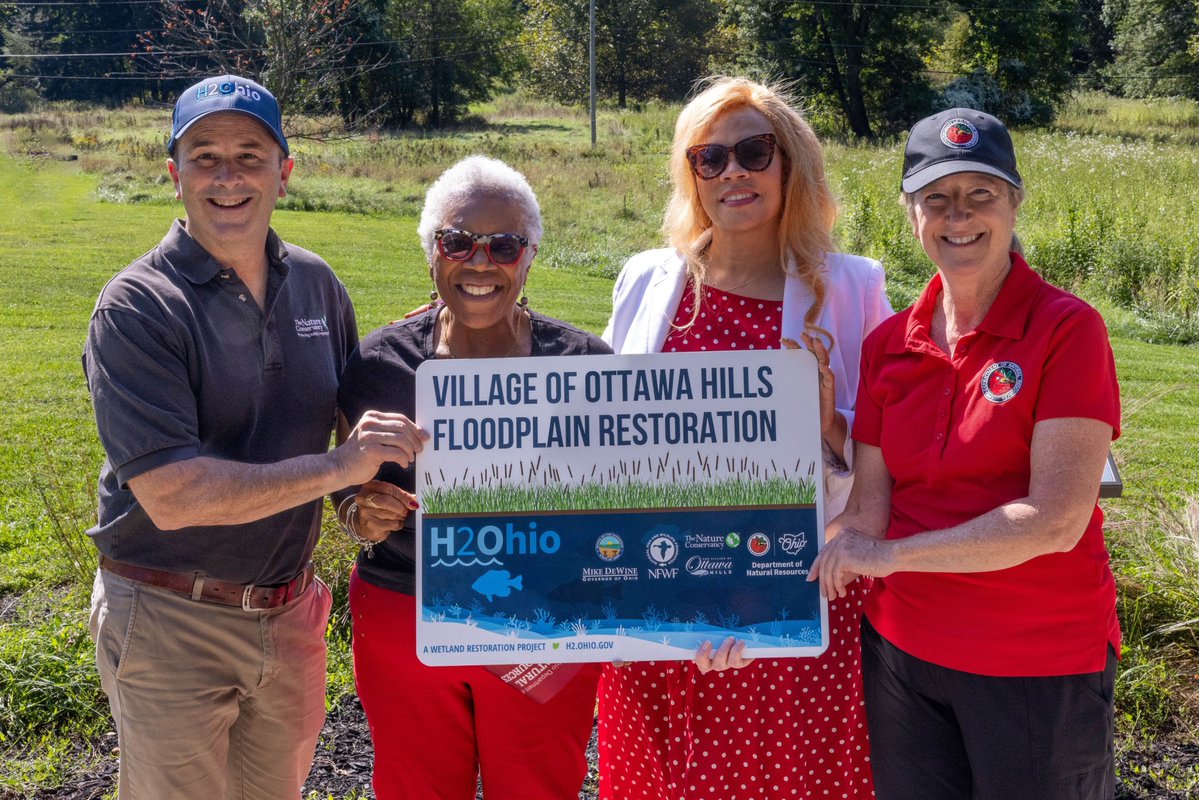 Four people are standing outdoors holding a sign that reads "Village of Ottawa Hills Floodplain Restoration" with logos of H2Ohio and other organizations. They are smiling in a grassy area with trees in the background.