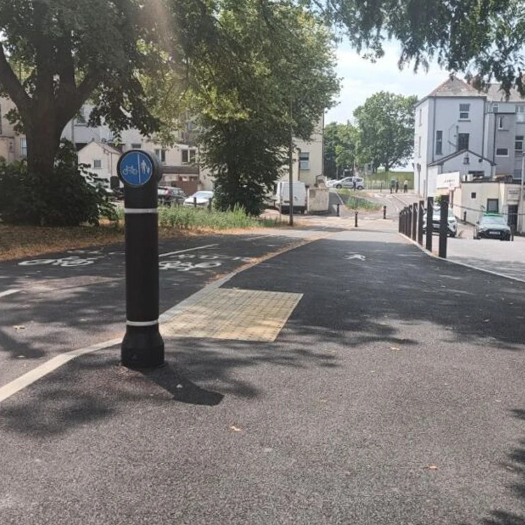 A paved shared pedestrian and cycle path slopes downhill through a leafy urban area. A black bollard with a blue cycling and walking sign stands in the foreground, beside tactile paving and marked cycle lanes. Trees shade the path, with buildings, parked vehicles, and a few people visible in the distance.