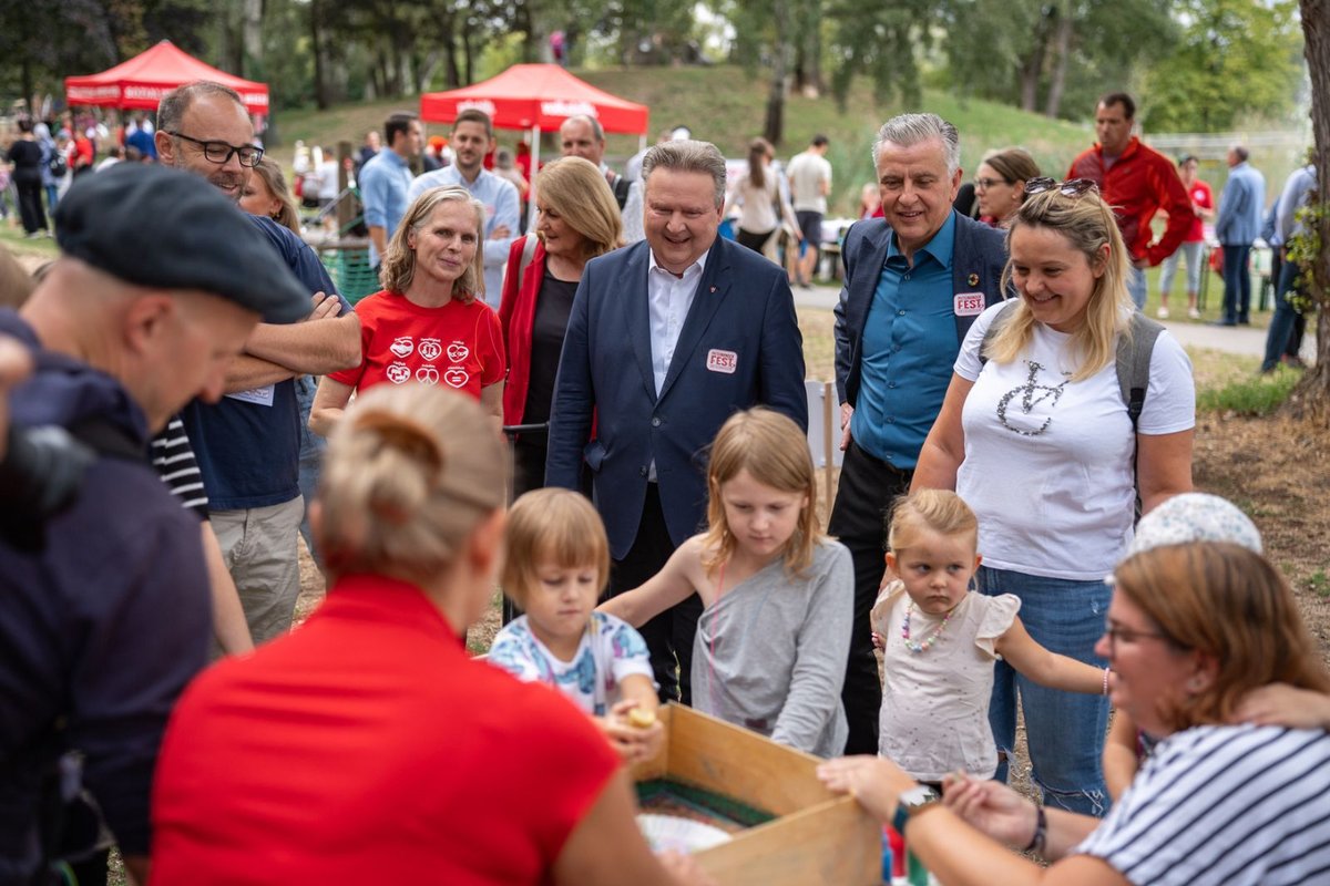 Eine Gruppe von Menschen, darunter Erwachsene und Kinder, steht unter roten Zelten in einem Park zusammen und unterhält sich.