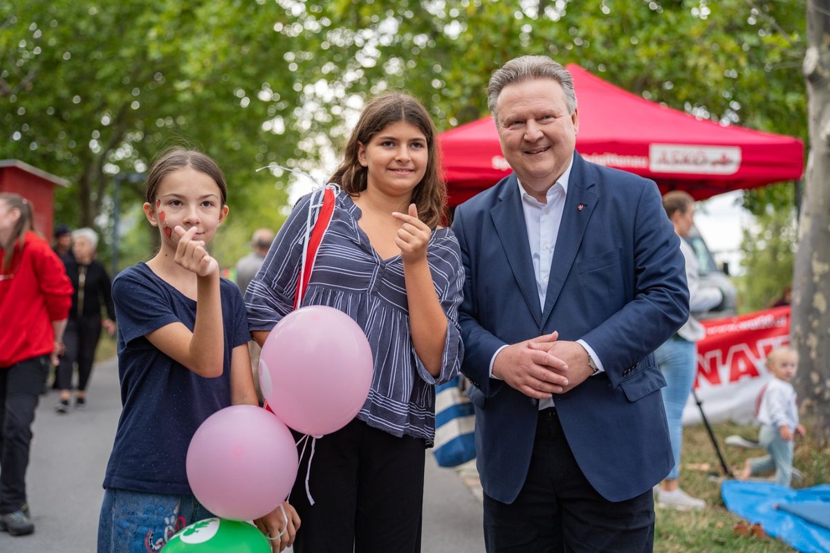 Drei Personen stehen zusammen auf einer Straße; ein Mann in einem blauen Anzug, eine Frau in einem gestreiften Oberteil und ein Mädchen in einem blauen T-Shirt, das zwei rosa Luftballons hält.