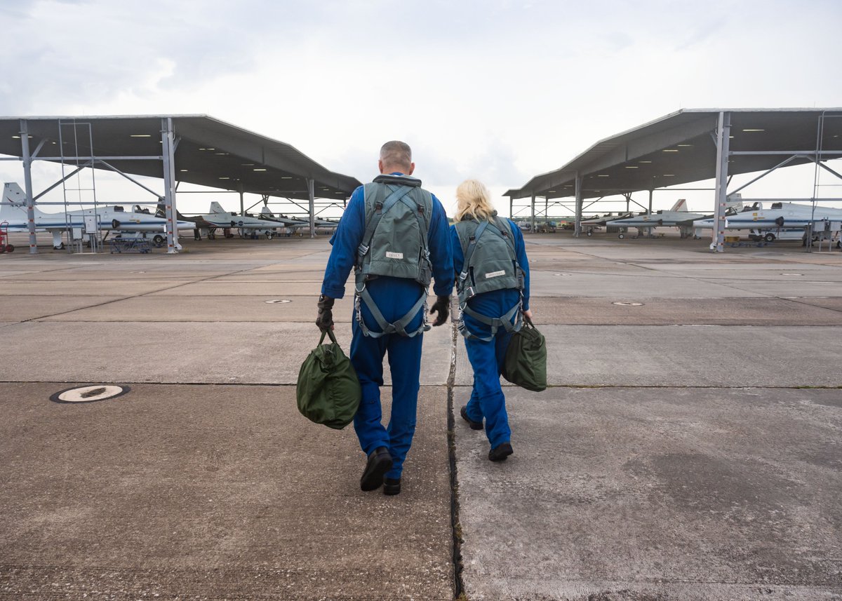 NASA astronauts Luke Delaney and Deniz Burnham walk toward their waiting T-38 aircraft ahead of a training flight. They each wear blue flight suits and harnesses and carry green duffle bags containing their helmets. Photo: NASA/Luna Posadas Nava