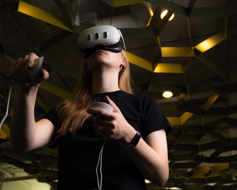 A student wearing a virtual reality headset holds two handheld controllers while standing beneath a geometric ceiling installation.