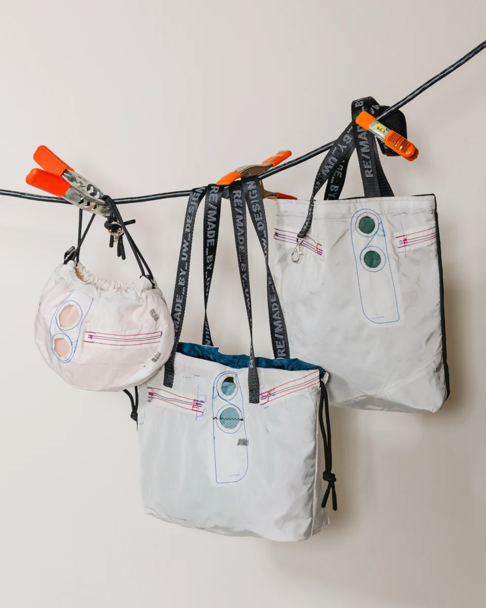 Three white, repurposed-fabric bags hang from a black wire with orange clamps. A small rounded bag, a medium tote, and a large tote feature blue and red stitching, circular details, and black straps with 'RE/MADE BY_UW_DESIGN' text.