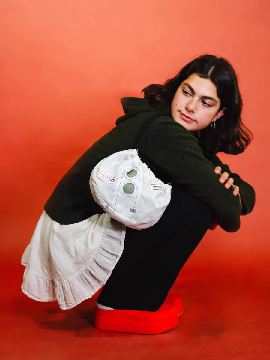 A young person squats, looking back over their left shoulder. They wear a dark green fleece, white ruffled garment, black pants, and red platform shoes. A white drawstring bag with blue & red stitching and two green circles hangs from their shoulder. The background is a solid reddish-orange.