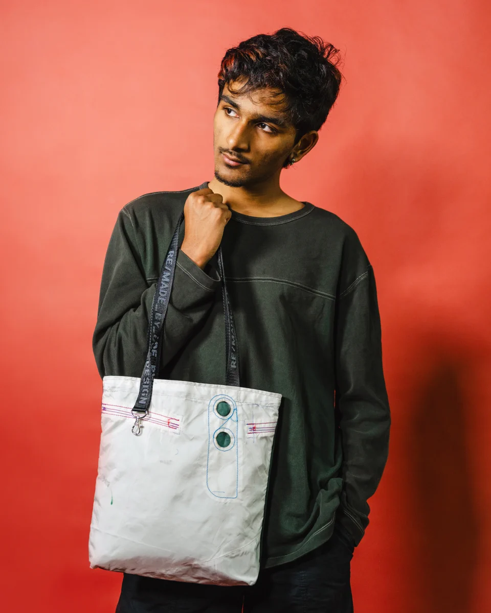 A young man with dark hair and reddish highlights, wearing a dark grey long-sleeved shirt, poses against a red background. He holds a white tote bag with blue stitching, red accents, and green grommets. The bag's strap reads \"RE/MADE BY SE DESIGN.\"