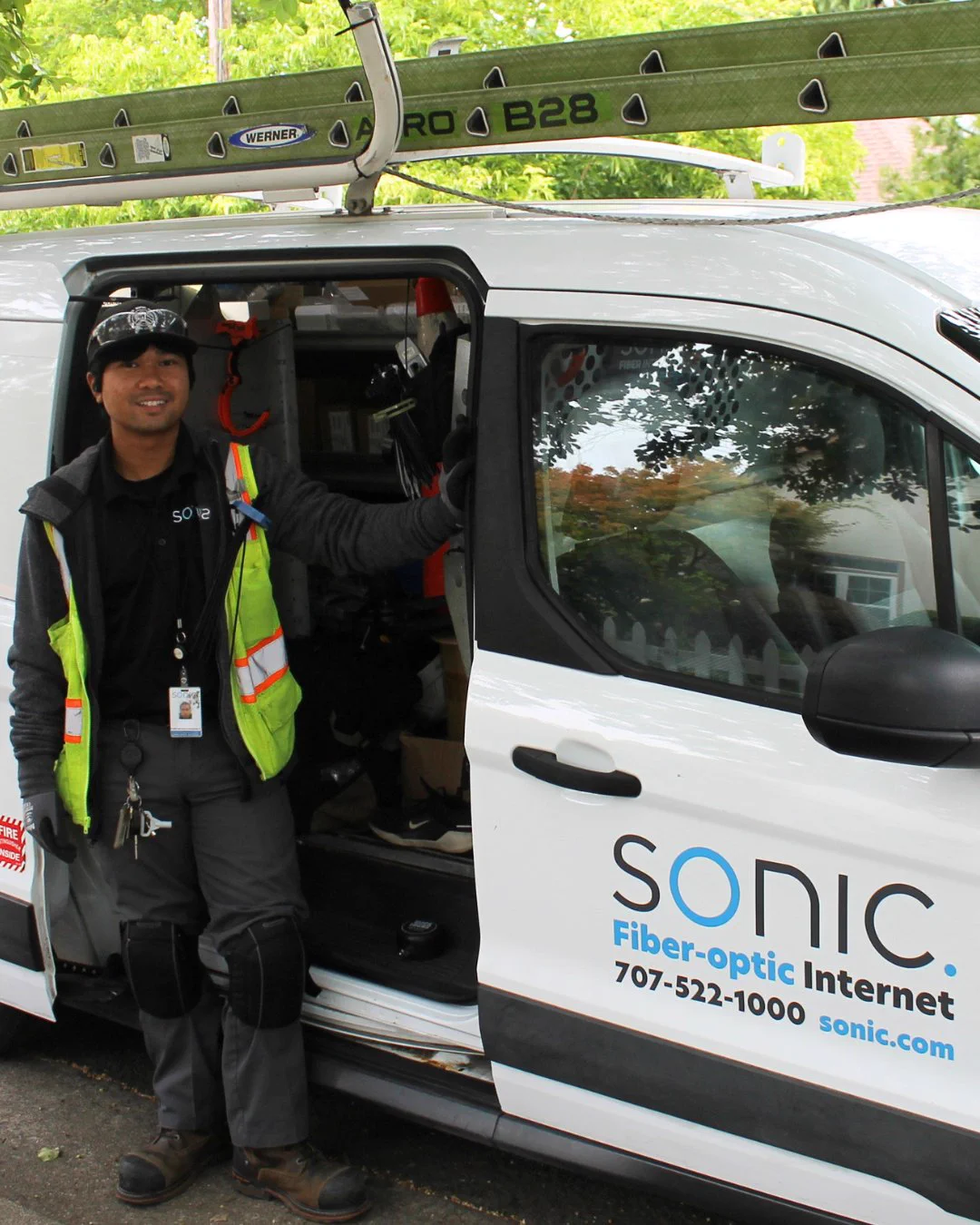 Randy, a Sonic Fiber Internet technician, stands and smiles beside an open white Sonic van parked under leafy trees in a residential neighborhood. A ladder is secured to the roof rack, and fiber installation equipment is visible inside the van. 