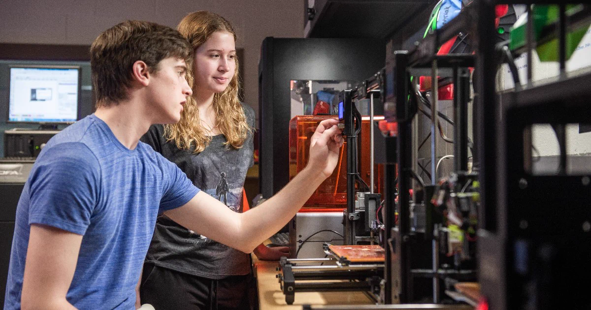 Two individuals are working with 3D printers in a lab. One is wearing a blue shirt and is adjusting a machine, while the other observes. A computer screen displaying technical information is seen in the background.