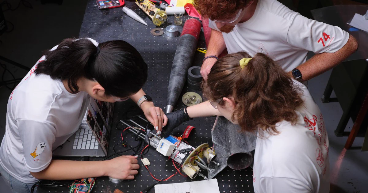 Three people work collaboratively on a technical project at a table covered with electronic components and tools. They focus on a device, surrounded by wires and a laptop. One person wears safety goggles.