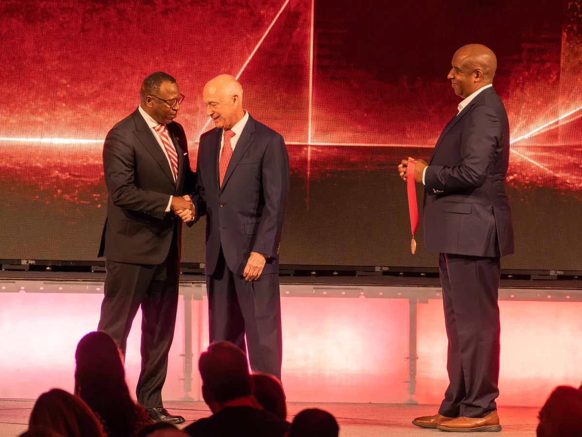 Two men in suits shaking hands on stage while a third man holds a red ribboned medal, all under dramatic red lighting.