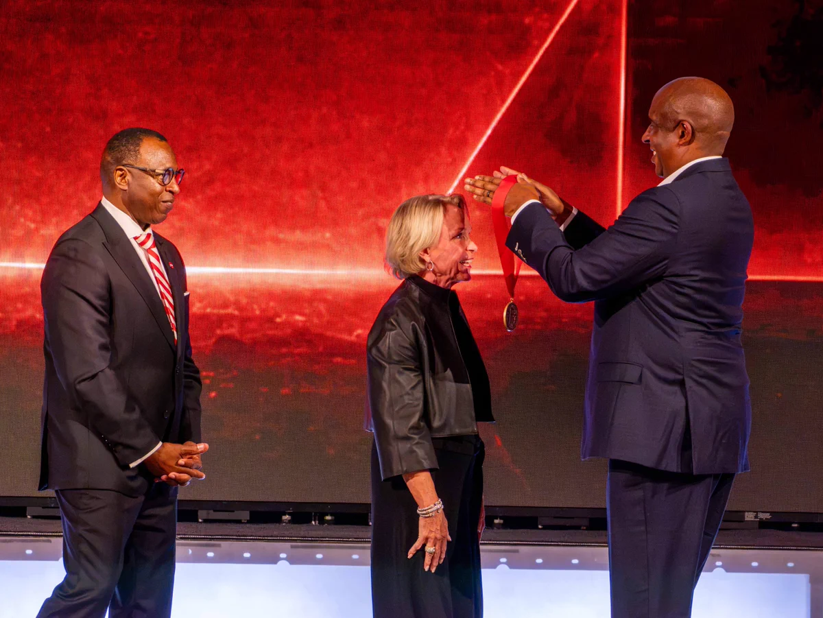 Man in a suit placing a medal around a woman's neck during an award ceremony with another man observing.