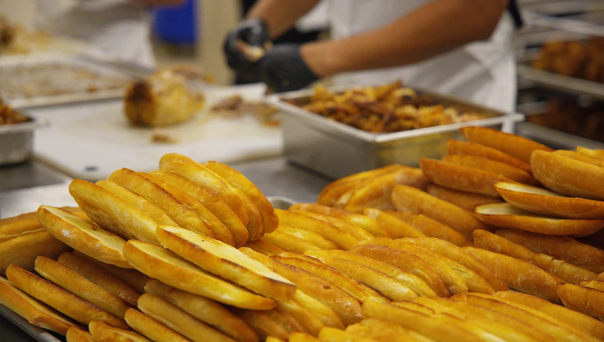 Stacks of freshly baked bread rolls are displayed on trays in a bakery setting. In the background, a person wearing gloves is preparing food at a countertop.
