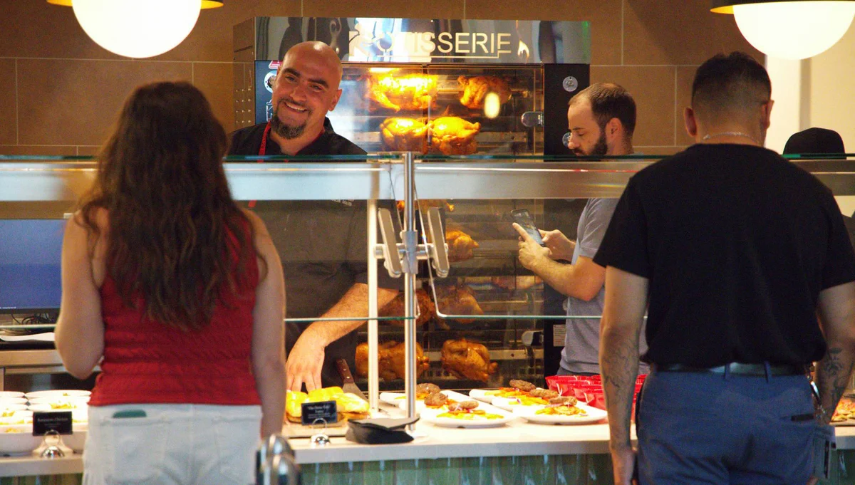 In a casual dining setting, three people stand at a counter with food displays. Behind the counter, a person smiles and interacts with the customers. Rotisserie chickens are visible in the background.