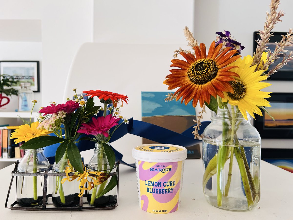 Photo of two flower arrangements with a pint of lemon curd blueberry made by Marcus ice cream in the middle and paintings and Slavo Cech sculpture behind.