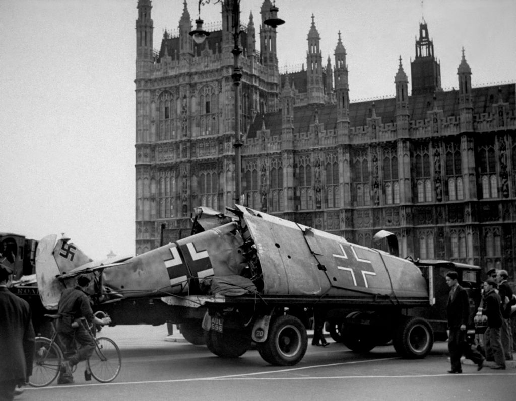 A damaged aircraft with swastikas on its tail and wings is being transported on a truck near the Houses of Parliament in London. Several people are gathered around the scene, and a cyclist is visible on the left. The architecture of the building serves as a backdrop.