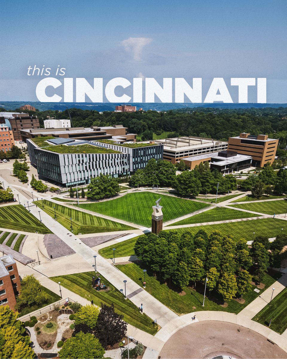 Aerial view of the University of Cincinnati campus with green lawns, walkways and academic buildings under a blue sky. Large white text reads, “this is CINCINNATI.”