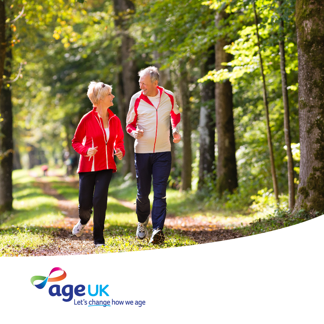 A person in a red jacket and another in a white and red jacket are jogging together on a forest trail. Lush green trees line the path. The logo of Age UK is visible in the lower part of the image, with the tagline "Let's change how we age."