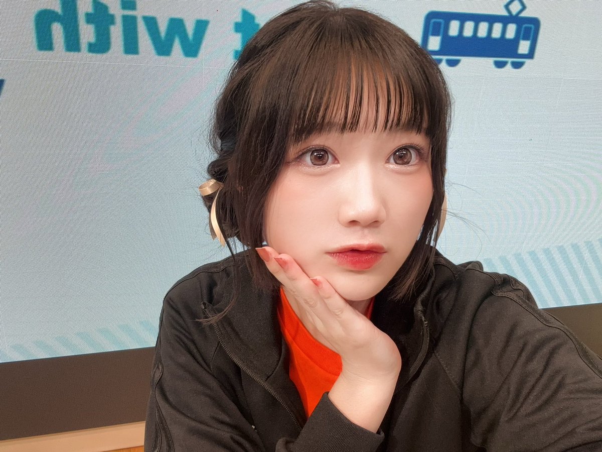 橘めいの自撮り写真です。
バストアップ画角です。右手を頬に当てています。
口は「う」の一歩手前です。カメラ目線です。

見えづらいですが、両サイドに低めお団子をしています。
黄色い細リボンをつけてもらいました！

黒いてつりょー会ジャージに、中にオレンジ色のてつりょー会Tシャツを着用しています。

背景は、てつりょーのアニメのタイトルロゴが映された電光掲示板です。