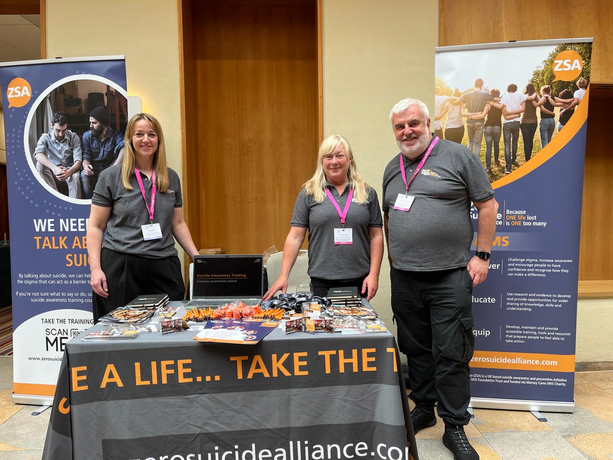 Three representatives stand behind a table with suicide prevention materials at an awareness event indoors.