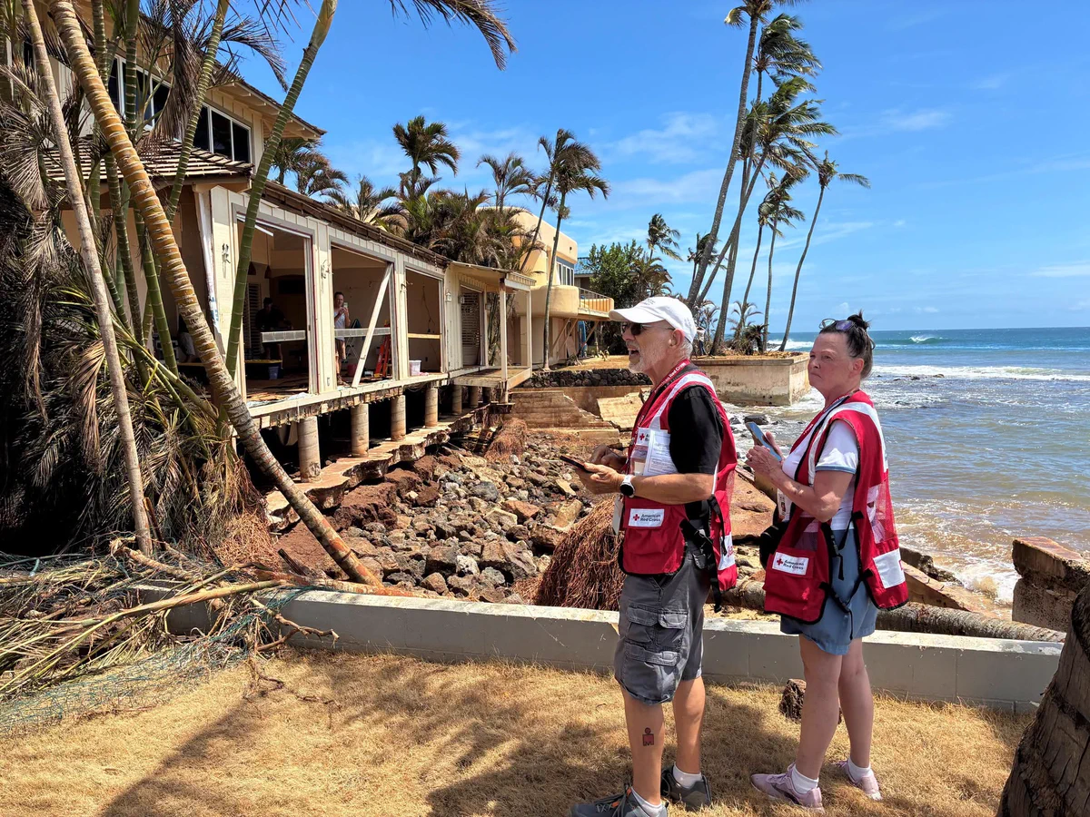 Two people in Red Cross vests assess damage near a coastal building with exposed foundations and fallen palm trees. The ocean and clear blue sky are visible in the background.