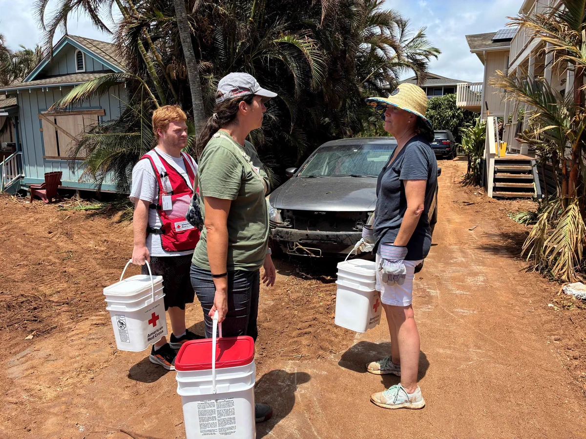 Three individuals are standing on a dirt path between houses, holding buckets. One person is wearing a vest with a Red Cross emblem. There's a car in the background surrounded by tropical plants. The group appears to be engaged in conversation.
