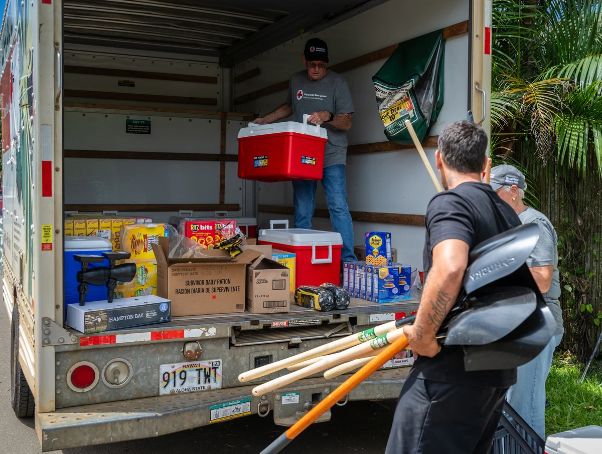 A person is inside a moving truck, organizing boxes of supplies, including bottled water and non-perishable food like Spam. Another individual stands nearby, holding shovels and rakes. The truck is parked on a tropical street with lush greenery. The truck's open door reveals a sign on the lower left referring to disaster relief.