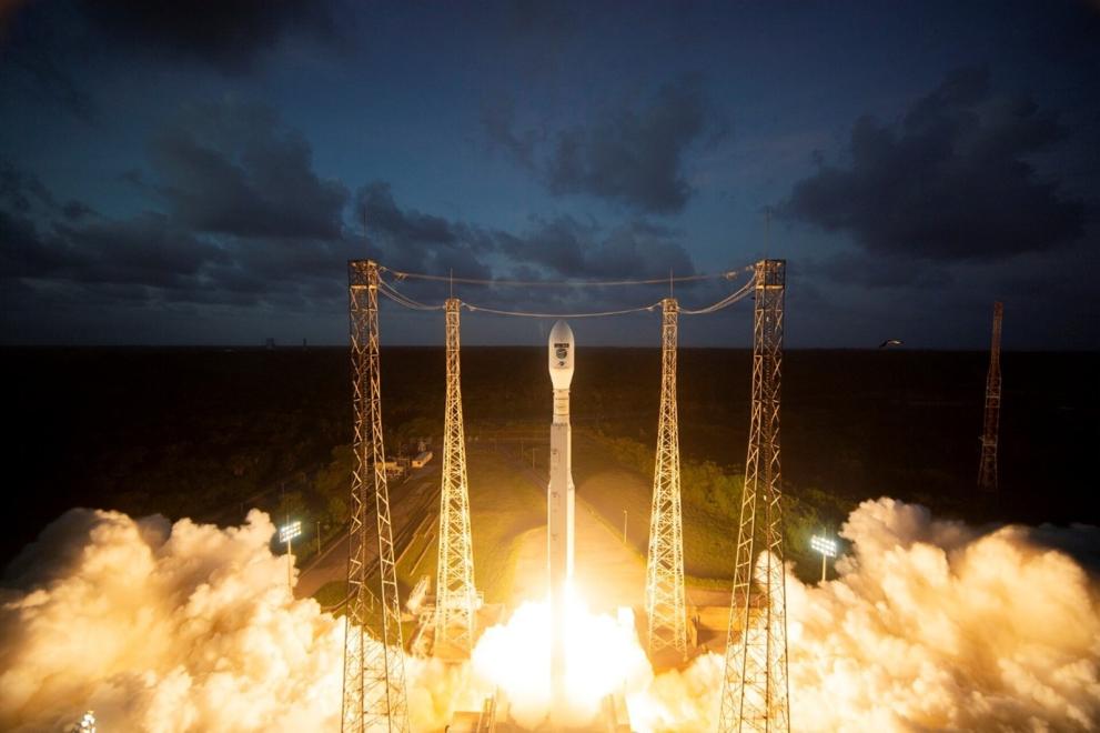 A rocket carrying the Copernicus Sentinel-3C satellite is launching into the night sky from the European Spaceport in French Guiana. Smoke and flames are visible beneath the rocket as it ascends. Tall structures surround the launch area. Dark clouds are visible above, contrasting with the illuminated area around the launch.