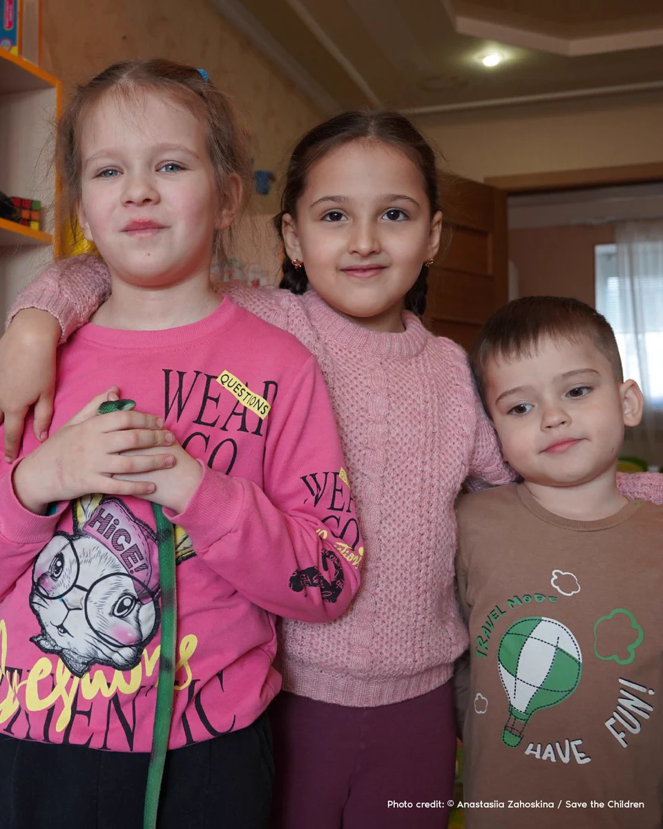 A young girl stands with her arms around another friend and her younger brother.