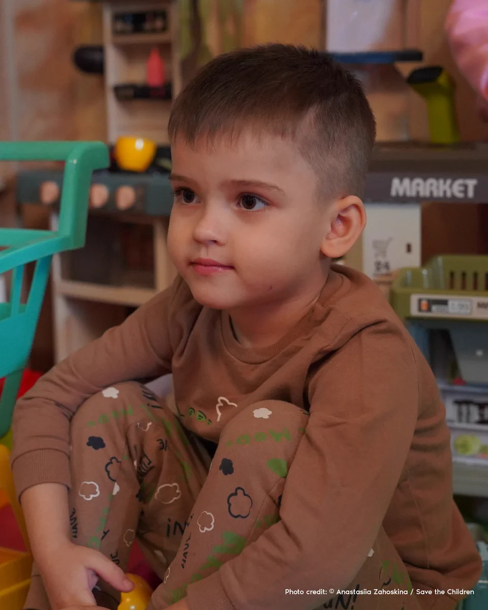 A young boy sits on the ground, his knees to his chest. A few toys can be seen around him in the background.
