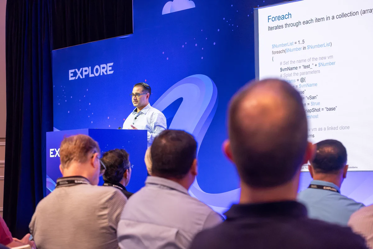 A person stands at a podium giving a presentation to an audience. The backdrop features the word "EXPLORE" prominently. The scene is from a conference or seminar setting.