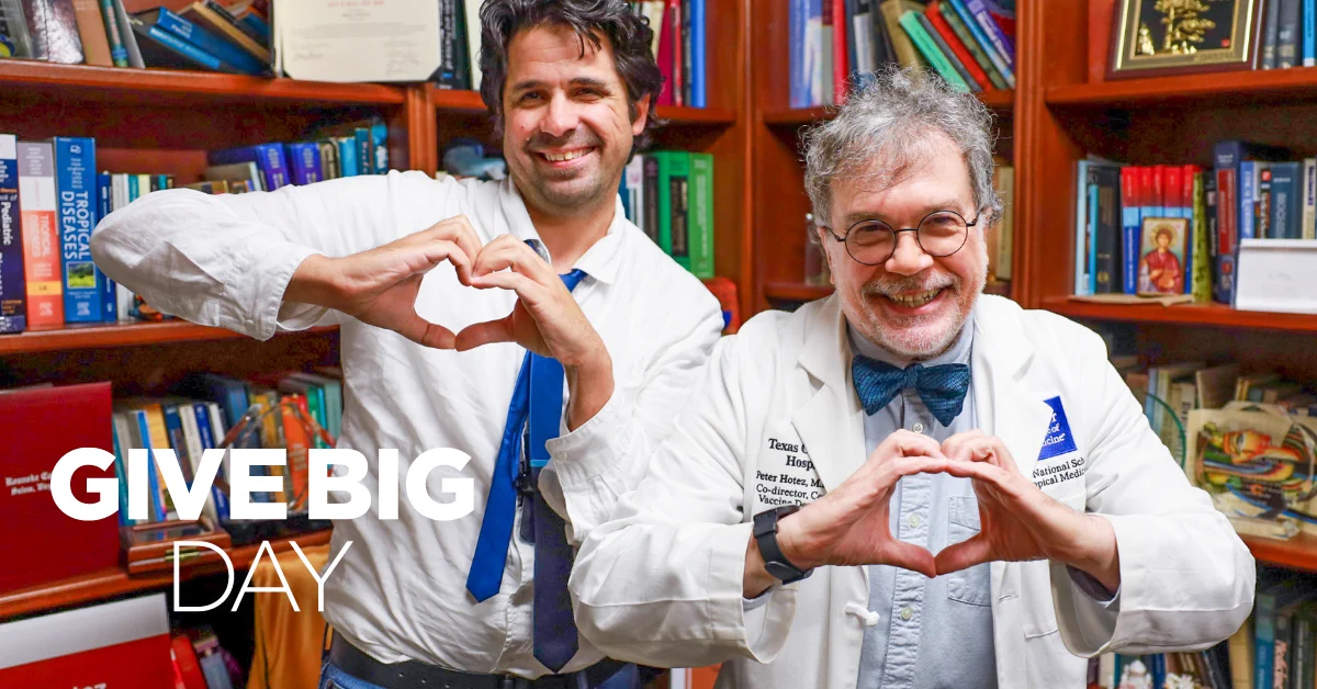 Two people stand in front of a bookshelf filled with books, making heart shapes with their hands. Both are smiling. Text on the image reads "GIVE BIG DAY." One person is wearing a white coat with a logo.