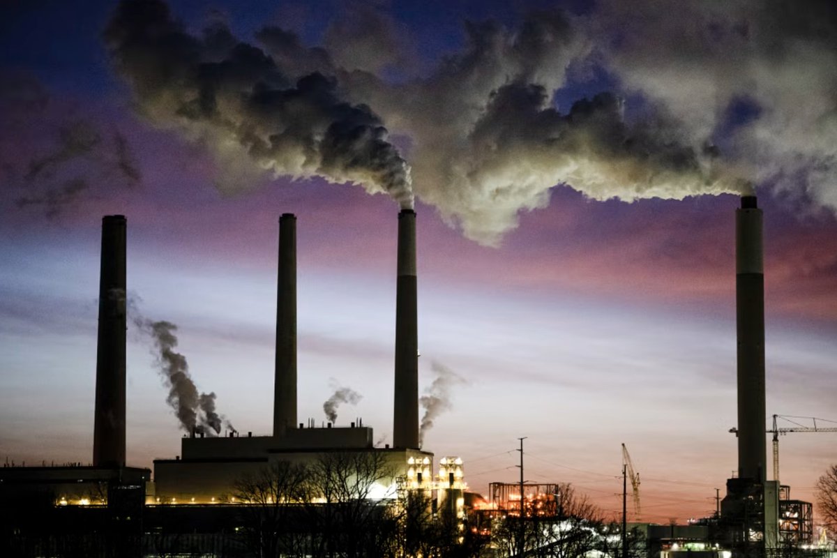 Smoke rises from tall industrial smokestacks against a twilight sky with purple and orange hues, with industrial facility lights glowing in the foreground